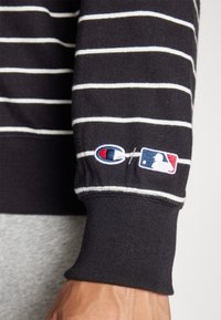 Fekete-fehér csíkos pulóver bordázott mandzsettával, hímzett Champion logóval és MLB logóval az ujjon. Puha pamut anyag.