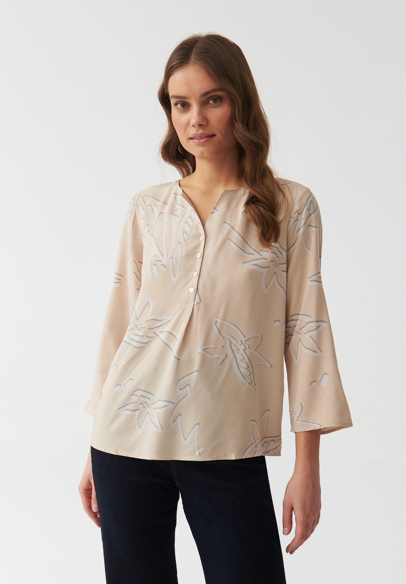 Hellbeige Bluse mit V-Ausschnitt, drei Viertel langen Ärmeln und grauem Blumenmuster. Weicher Stoff mit Knopfdetails am Ausschnitt.