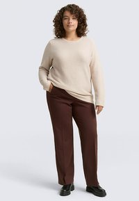 Lichtbeige trui met textuur en een ontspannen pasvorm, gecombineerd met bruine broek met rechte pijpen en zwarte schoenen. Model staat met handen in de zakken.