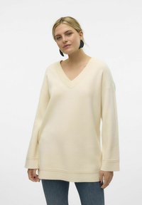 Pull oversized crème avec un col en V, un col côtelé, des épaules tombantes et des manches longues. Il présente un ourlet droit et une texture douce.