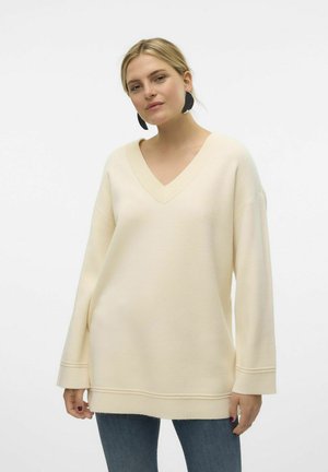 Cream-colored oversized sweater with a V-neck, ribbed neckline, dropped shoulders, and long sleeves. Features a straight hem and soft texture.