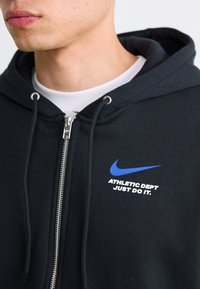 Nærbillede af en person iført en sort zip-hættejakke med et blåt Nike swoosh og teksten "ATHLETIC DEPT JUST DO IT." på brystet.