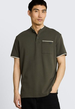 Junger Mann trägt dunkelgrünes kurzärmeliges Polo mit beiger Blende und Brusttasche, steht mit einer Hand in der schwarzen Hosentasche.