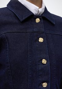 Veste en denim bleu foncé avec des boutons dorés en forme de rose portée sur une chemise blanche à col.