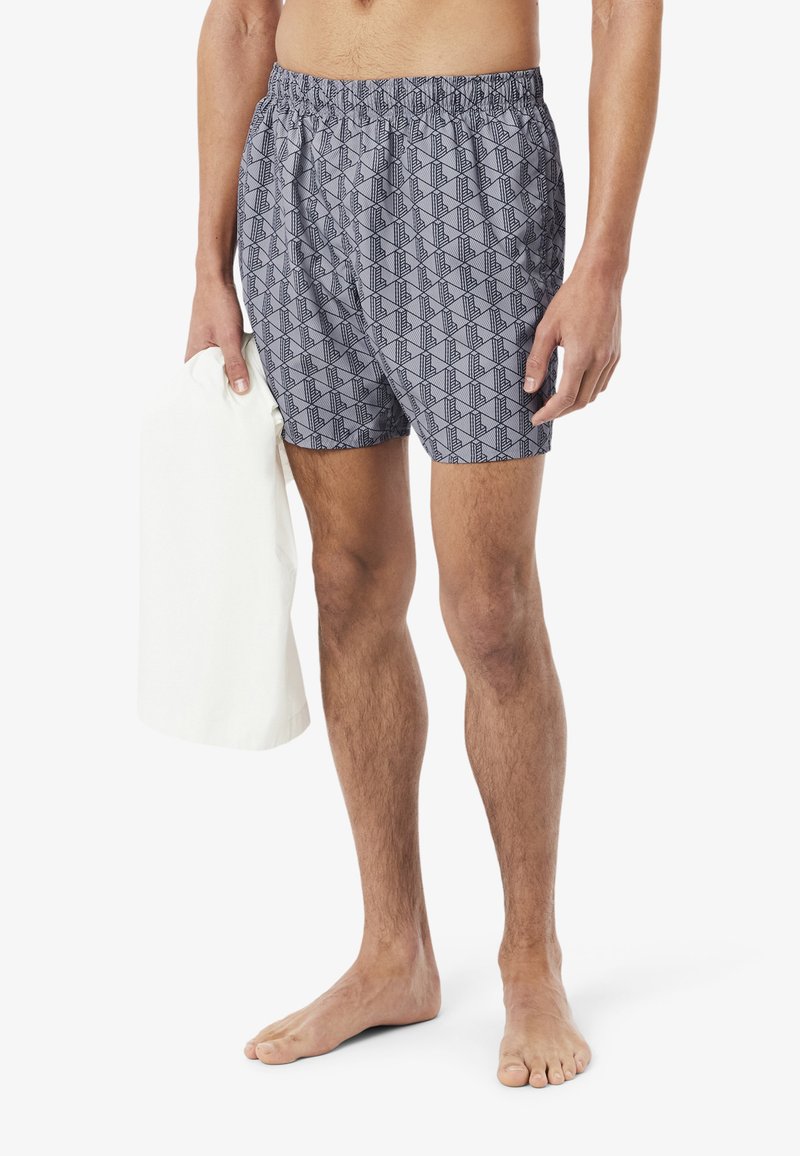Modèle masculin portant un short de bain gris à motifs, tenant une serviette blanche dans une main, debout pieds nus sur un fond blanc.