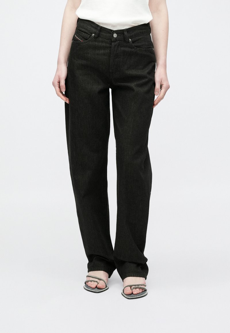 Žena oblečená v čiernych džínsach s rovnými nohavicami a otvorenými sandálmi na vysokom podpätku, stojaca pred jednoduchým bielym pozadím.