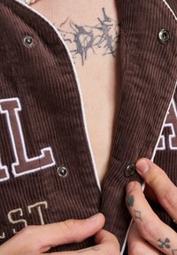 Indumento in velluto a coste marrone con piping bianco, caratterizzato da bottoni a pressione argentati e scritte ricamate. Tatuaggi visibili sulla mano e sul petto.