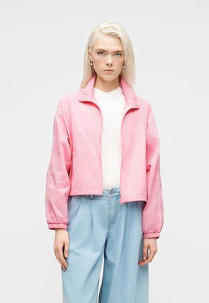 Donna bionda che indossa una giacca rosa chiaro con zip sopra una maglietta bianca, abbinata a jeans azzurri larghi, in piedi davanti a uno sfondo semplice.