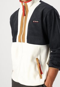 Giacca in pile con pannelli neri, crema e marroni. Colletto con cerniera, tasche laterali con accento rosso e design a blocchi di colore distinto.