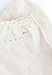 Beige bomullsshorts med elastisk midja, platt bakficka och en liten broderad orange logotyp på vänster sida.