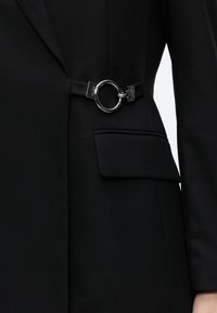 Personne portant un blazer noir avec une poche à rabat et un détail anneau argenté à la taille ; seuls le torse et une partie du bras sont visibles.