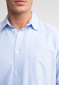 Mann trägt hellblaues Hemd mit Brusttasche, teilweise geknöpftem Kragen vor neutralem Hintergrund.