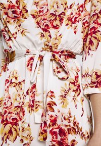 Robe portefeuille florale avec un fond blanc, de grands motifs de roses rouges et jaunes, une ceinture nouée à la taille en tissu doux et des manches froncées.
