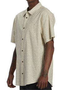 Korte mouw knoopshirt in lichtbeige met een subtiel geometrisch patroon, voorzien van een borstzak en bruine knopen. Zachte textuur.