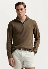 Jeune homme portant un pull marron à zip quart, un short blanc, une ceinture marron et une montre au poignet, se tenant devant un fond uni.