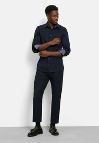 Marineblaues Hemd mit Knöpfen und gemusterten Manschetten, kombiniert mit dunklen Hosen und schwarzen Schuhen. Glattes Material mit klassischem Kragen und taillierter Passform.