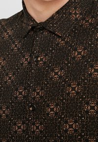 Camicia nera con bottoni caratterizzata da un motivo floreale in tonalità rame, tessuto testurizzato e design del colletto classico. Un bottone visibile.