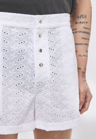 White embroidered shorts featuring floral patterns and cut-outs, a front button closure with metal buttons, and a straight hem.