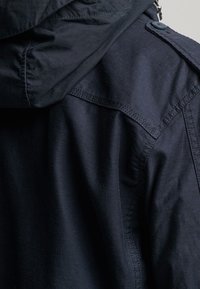 Donkerblauwe jas met capuchon, met een gladde textuur, duidelijk stikwerk en metalen accenten bij de schouder en kraag.