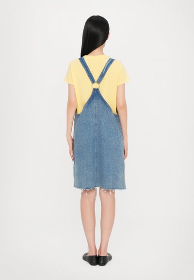 Robe en salopette en denim avec un ourlet effiloché, des bretelles ajustables et un détail en métal circulaire, associée à un t-shirt jaune.