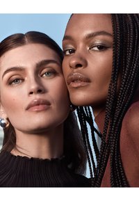 Zwei Nahaufnahmen von Frauen mit unterschiedlichen Hauttönen. Eine hat lange dunkle Haare in Zöpfen, die andere hat glattes braunes Haar und trägt ein schwarzes, geripptes Oberteil.