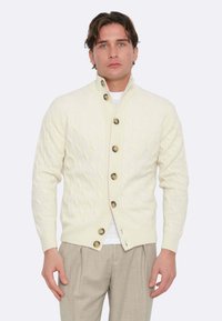 Cardigan lavorato a maglia crema con colletto alto, caratterizzato da grandi bottoni marroni. Tessuto morbido, design aderente, abbinato a pantaloni chiari.