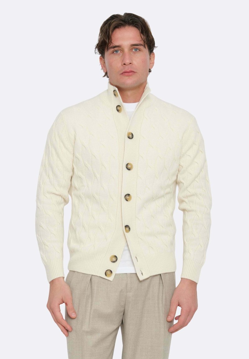 Cardigan lavorato a maglia crema con colletto alto, caratterizzato da grandi bottoni marroni. Tessuto morbido, design aderente, abbinato a pantaloni chiari.