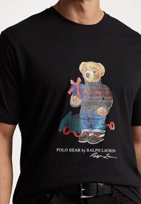 Medvěd vesele oblečený do svátečního svetru, džínů a mokasín, držící dárkovou krabičku, pod nímž jsou další zabalené dárky a červená stuha.