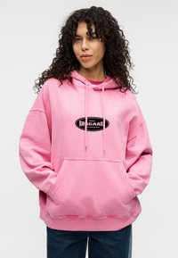 Ροζ φαρδιά φούτερ με καγκουρό τσέπη μπροστά και κουκούλα με κορδόνια. Διαθέτει μαύρο οβάλ λογότυπο στο κέντρο. Μαλακή υφή υφάσματος.