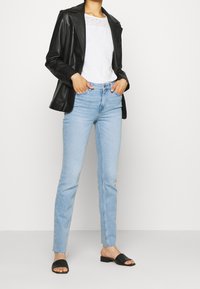 Blazer de cuero negro sobre una camisa blanca con detalles de ojal, combinado con jeans de color azul claro y sandalias negras tipo slide.