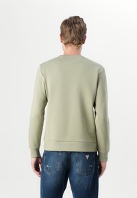 Sudadera verde oliva claro con un cuello redondo, puños y dobladillo acanalados, textura suave, usada sobre unos vaqueros azules. Etiqueta de logo visible en los vaqueros.