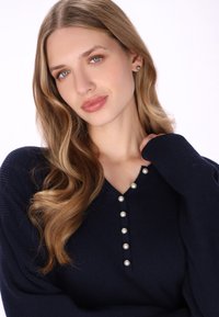 Pulover tricotat cu nervuri, de culoare bleumarin, cu decolteu în V și șapte nasturi de sidef pe partea frontală. Mâneci lungi cu o croială ușor supradimensionată. Textură moale.