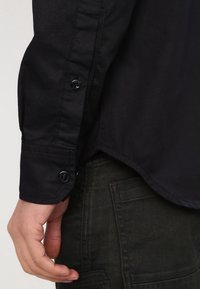 Närbild av en persons vänstra hand som vilar bredvid fickan på mörk denimjeans, iförd en svart långärmad skjorta med knäppta manschetter.