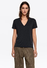 Czarny T-shirt z dekoltem w serek, krótkimi rękawami i subtelnymi zdobieniami w postaci ćwieków ułożonych w wzór. Wykonany z miękkiej tkaniny. Połączony z wzorzystymi, luźnymi spodniami.