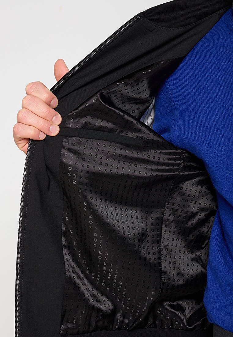 Schwarze Jacke mit einem glatten schwarzen Innenfutter, das ein dezentes eingeprägtes Logomuster aufweist. Sichtbarer Reißverschluss und gerippte Kragen Details.