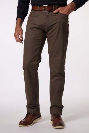Mann trägt braune Hose, braunen Ledergürtel, dunkles Langarmshirt und braune Lederschuhe mit hellen Sohlen, steht mit einer Hand in der Tasche.