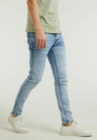 Vaqueros ajustados de mezclilla azul claro, con un sutil desgaste y patrones de arrugas, combinados con zapatillas blancas, mostrando una vista lateral.