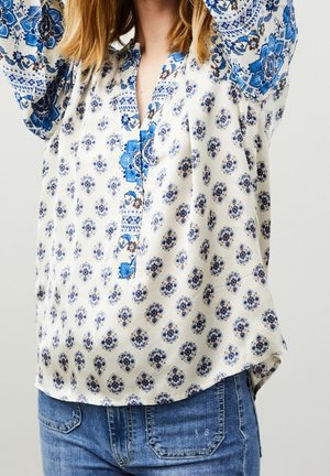 Blusa in satin con un motivo floreale blu su sfondo crema, dotata di colletto a V e maniche lunghe, arricchita da un orlo curvo.