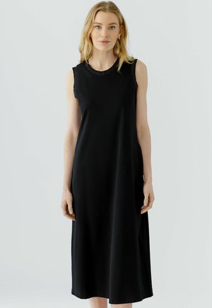 Femme blonde portant une robe midi noire sans manches avec un col rond et une bordure texturée, debout devant un fond uni clair.