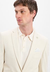 Hellbeige Blazer aus strukturiertem Stoff mit Revers, kombiniert mit einem passenden beigen Poloshirt mit Drei-Knopf-Verschluss und weißem Einstecktuch.