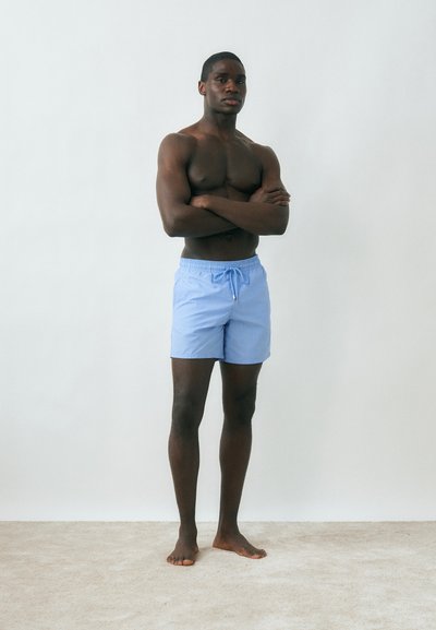 Μυώδης άνδρας όρθιος ξυπόλυτος με σταυρωμένα χέρια, φοράει ανοιχτό μπλε μαγιό μπροστά σε απλό λευκό φόντο και μπεζ πάτωμα.