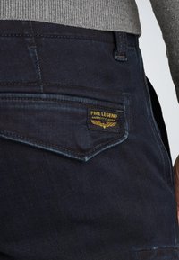 Donkerdenim jeans met een slim fit, voorzien van een achterzaklabel "PME Legend American Classic" in goud op een zwarte achtergrond.