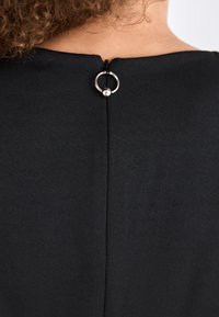 Černý oděv s kulatým stříbrným knoflíkovým zapínáním na zádech, hladká tkanina a jemný svislý šev.
