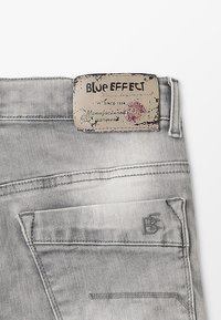 Jeans de denim gris con una superficie texturizada, que presentan una etiqueta de cuero con el logo "BLUE EFFECT" en la cinturilla y un detalle en el bolsillo trasero.