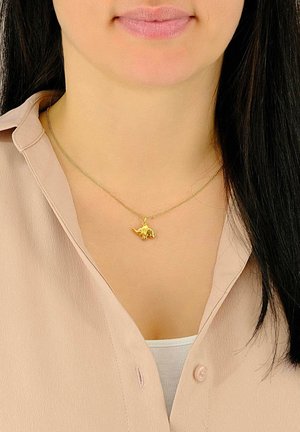 Femme aux longs cheveux foncés portant un chemisier beige clair et un collier avec un pendentif en forme de dinosaure en or sur une fine chaîne.