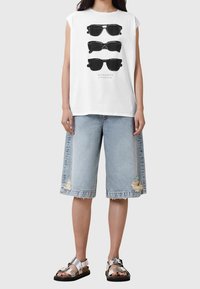 Camiseta sin mangas blanca con un gráfico negro de tres gafas de sol, combinada con shorts de mezclilla azul claro con dobladillos deshilachados y rotos. Sandalias plateadas.