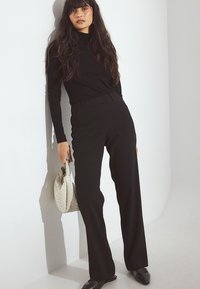 Melnā pieguļošā augstais kakls, kas kombinēts ar augsto jostasvietu melnajām biksēm. Modelis tur rokās austu baltu rokassomu un valkā melnus sausu purngalu apavus.