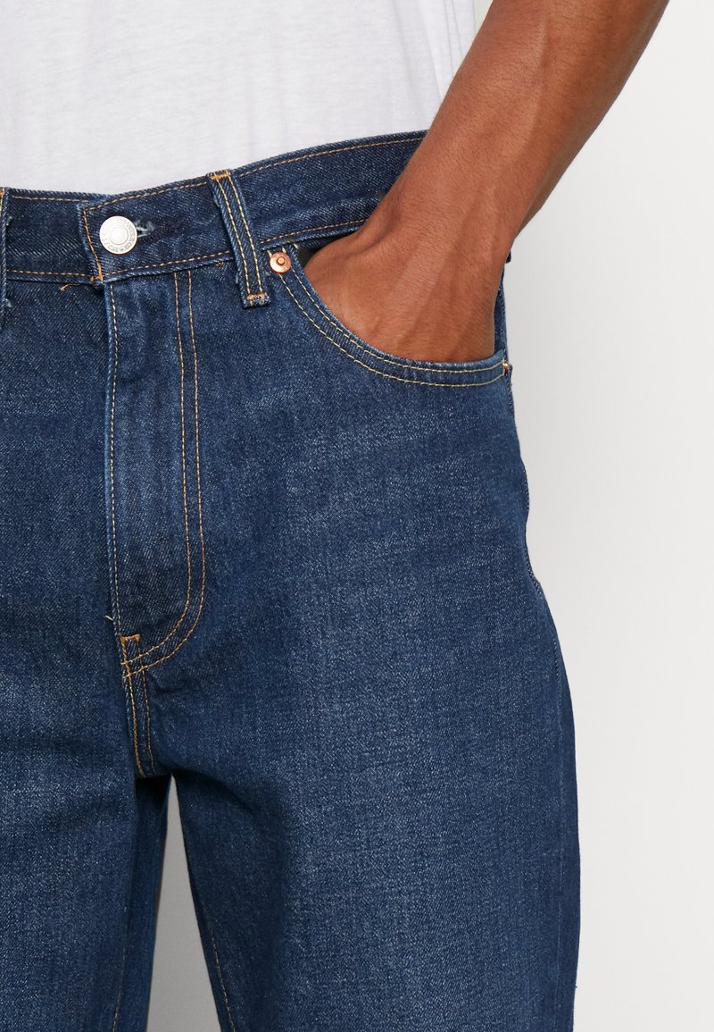 Jeans in denim blu scuro con design a gamba dritta, caratterizzati da rivetti in ottone, cuciture arancioni e una tasca frontale. Il materiale sembra robusto e texturizzato.