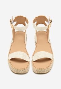 Beige espadrillesandaler med flettet stroppedesign, spænde lukning og en tekstureret sål. Har en flad, åben tå-stil med synlige syninger.