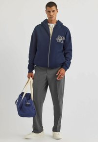 Granatowy rozpinany kaptur z logo z przodu, szare dopasowane spodnie i niebieska torba sportowa. Białe trampki dopełniają stylizację.